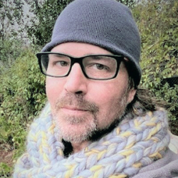Man wearing glasses, a knit beanie, and a chunky scarf stands outdoors with greenery in the background, reflecting on how to heal through art and the calming influence of nature.