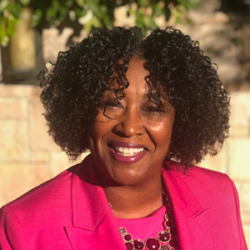 Smiling woman with curly hair in a pink blazer standing outdoors in sunlight, embodying confidence and resilience—qualities essential for those learning how to lead during a disaster and racial reckoning.