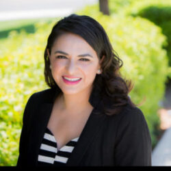 Woman with dark hair in a black blazer smiling outdoors, green bushes behind her—she embodies confidence and knows how to advocate for others in every setting.