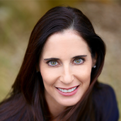 Woman with long brown hair and green eyes smiling at the camera, outdoors with a blurred background—her calm presence contrasts the typical stress seen during communication failures in disaster situations.