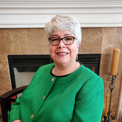 Smiling woman with short gray hair and glasses, wearing a bright green jacket, sitting indoors by a fireplace, discusses communication failures in disaster response.