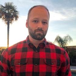 Man in a red plaid shirt stands outdoors at sunset with palm trees in the background, reflecting on how to create a rebuild organization that inspires positive change.