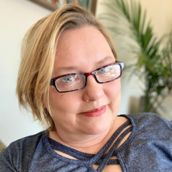 Woman with short blonde hair and glasses, wearing a blue top, smiling indoors with a plant in the background—exploring how to Block Captain for her local community.