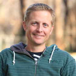 Smiling man in a green hoodie sitting outdoors with a blurred background of trees, perhaps reflecting on How to Create a Rebuild Organization.