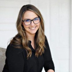 Woman with long brown hair and glasses, wearing a black top, smiling while seated against a light background—perfect for sharing tips on How to Zone Captain: Rural Area.