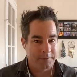 Man with dark hair and earphones sits indoors, a go bag visible beside him, with photos and hanging decor on the wall behind—a subtle nod to how to prepare for disaster.