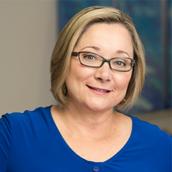 Woman with short blonde hair, glasses, and a blue top, smiling at the camera in an indoor setting—perhaps ready to share insights on how to create a mental health system.