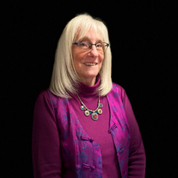 Smiling older woman with straight gray hair, glasses, and a purple top and vest, posing against a black background—Margie Foster from "How to Lead a Community Post Disaster.