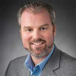Smiling man with short hair and a beard, wearing a gray suit jacket and blue shirt, on a dark background, featured in How to Lead: Public Sector (Local) with James Gore.