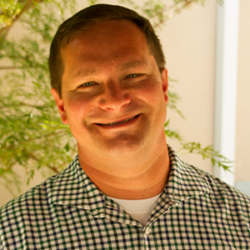 Smiling man in a checkered shirt stands in front of greenery and a beige wall, facing the camera—perfect image for discussing How to Manage Private Lands in the Era of Mega Fires with Algeo Che Casul.