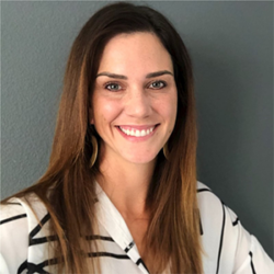 Woman with long brown hair, smiling, wearing a white blouse with black stripes, standing by a gray wall, ready to share insights on How to Design and Implement a Mental Health Collaborative with Adrienne Heinz.