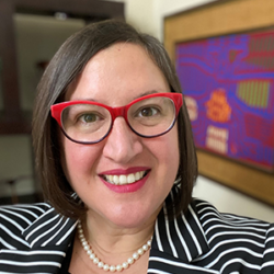 Smiling woman with short brown hair, red glasses, striped blazer, and pearl necklace indoors, featured in Episode 9: How to Serve Communities Pre and Post Disaster with Christa López PhD CEM.