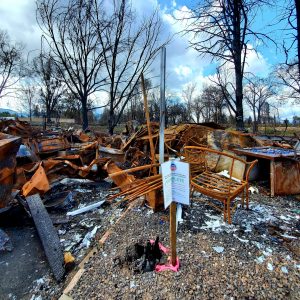 scene of a home destroyed by wildfire under a blue sky, with a jumbled pile of rusted, burned and mangled debris and barren tree limbs behind