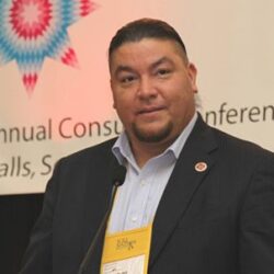 Man in a suit speaking at a conference, standing at a podium with a microphone and event sign behind him, addressing national leaders and the Board of Directors on challenges faced by fire affected communities.