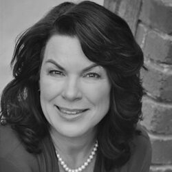 Smiling woman with wavy dark hair, pearl necklace, and blazer, posing in front of a brick wall—an inspiring member of the Board of Directors supporting fire affected communities.