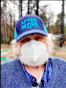 close up photo of a white haired man with blue shirt and blue cap with After the Fire printed on it