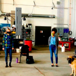 Six people and a dog, all spaced apart and wearing masks, stand in a large industrial workshop—wildfire survivors gathering to support community rebuild efforts.