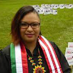 A graduate wearing glasses and a colorful sash stands outside on grass with signs in the background, embodying the spirit of "How to be an Emergent Leader with Jocksana Corona.