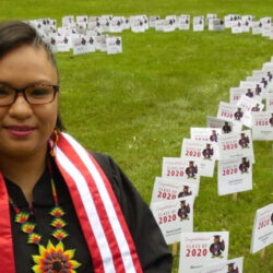 A graduate in a gown stands on grass near a display of small Class of 2020 signs, celebrating achievement and reflecting on lessons like How to be an Emergent Leader with Jocksana Corona.