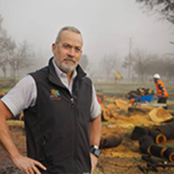 A man stands in front of cut logs, with workers and foggy trees in the background, reflecting on how to rebuild as a public sector official with Tennis Wick and navigating the rebuild permit process.