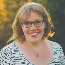 Smiling woman with glasses and wavy hair outdoors, wearing a striped shirt and necklace—Amy Simpkins enjoys the fresh air while discussing a rebuild permit.