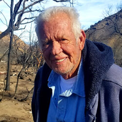 Elderly man with white hair, identified as Gary Jones, stands outdoors in a burned, barren landscape with charred trees, contemplating the next steps as he awaits a rebuild permit.
