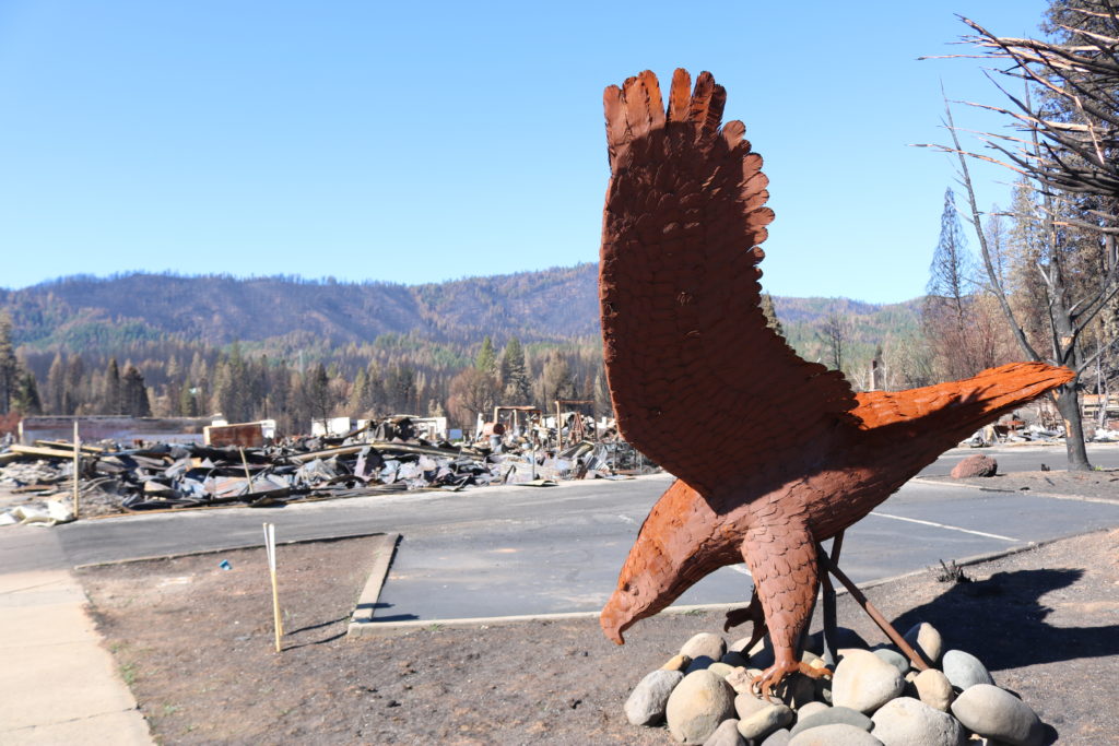 Greenville, CA view of downtown after Dixie Fire with large fire-tinged rusted metal sculpture of an eagle in the foreground.