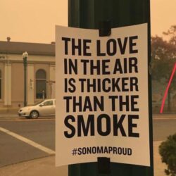 A sign reads The love in the air is thicker than the smoke. #SonomaProud, offering a moment of remembrance and reflection on a smoky street.
