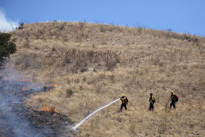 Three firefighters on a dry grass hill spraying down a burned patch of grass