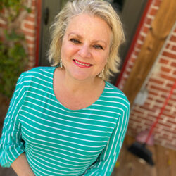 Smiling woman with blonde hair, Cherry Jochum, in a green and white striped shirt stands outside by a brick wall.