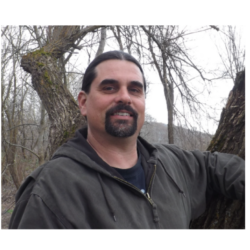 Man with dark hair and beard standing outdoors by a tree, wearing a brown jacket, with bare trees in the background, embodying resilience amidst nature’s quiet strength.