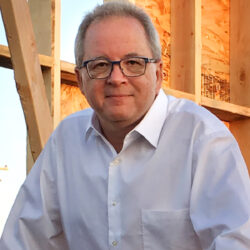 A man in glasses and a white shirt stands in front of a wooden construction frame outdoors, perhaps awaiting a rebuild permit, reminiscent of Brian Fies’s journey after loss.