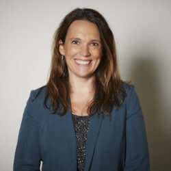 Renae Hanvin, with long brown hair in a blue blazer, smiles against a plain light-colored background, ready to discuss your rebuild permit needs.