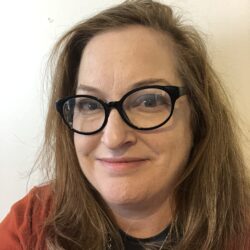 Heather Milton, with wavy light brown hair, wearing black glasses and a red top, smiles at the camera as she discusses the rebuild permit.