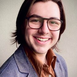 Jacob Bentz, a young man with shoulder-length brown hair, glasses, and a gray blazer, smiles at the camera against a plain background—ready to discuss your rebuild permit needs.