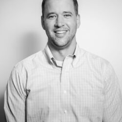 Smiling man, Trey Bearden, in a light plaid shirt stands against a plain background, facing the camera.