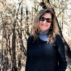 Woman with wavy hair and sunglasses smiling in a sunlit forest, wearing a dark sweater and gray scarf, radiates the warmth of community amidst nature’s artful beauty.