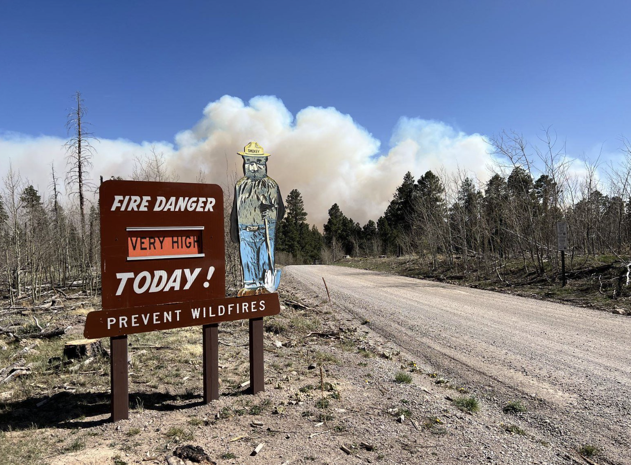 daytime picture of a state park sign with high fire danger words and smoke plumes in the background
