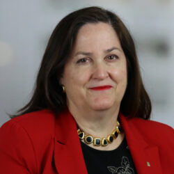 A woman with long brown hair, red lipstick, and a red blazer, wearing a gold and black necklace, smiles—much like Katherine Hammack at a rebuild permit celebration.