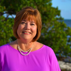 Smiling woman, Michelle Luckett, in a purple top and pearls, standing outdoors near water and green trees.