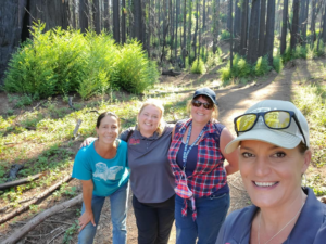 four women standing together smiling with a forested area behind