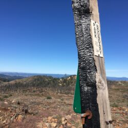 A burned wooden post with a Park Boundary sign stands in the rocky, open landscape of Hood Mountain under a clear blue sky, evidence of recent prescribed burning.