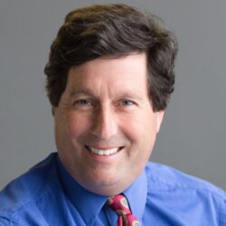 Smiling man with dark hair, Louie Delaware, wears a blue shirt and red paisley tie, posed against a gray background—ready to answer your rebuild permit questions.