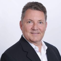 Smiling man, possibly Steve Crout, in a black suit jacket and white shirt, facing forward against a plain white background—ready to discuss your rebuild permit needs.