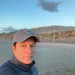 Man in a jacket and cap taking a selfie on a sandy beach with hills and a cloudy sky in the background, capturing the spot where Stuart Mitchell once awaited his rebuild permit.