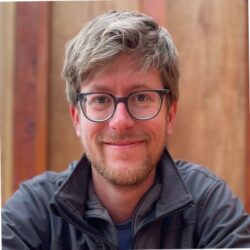 A man with glasses and a beard, possibly Will Heegaard, smiles at the camera while sitting in front of a wooden background, perhaps discussing a rebuild permit.