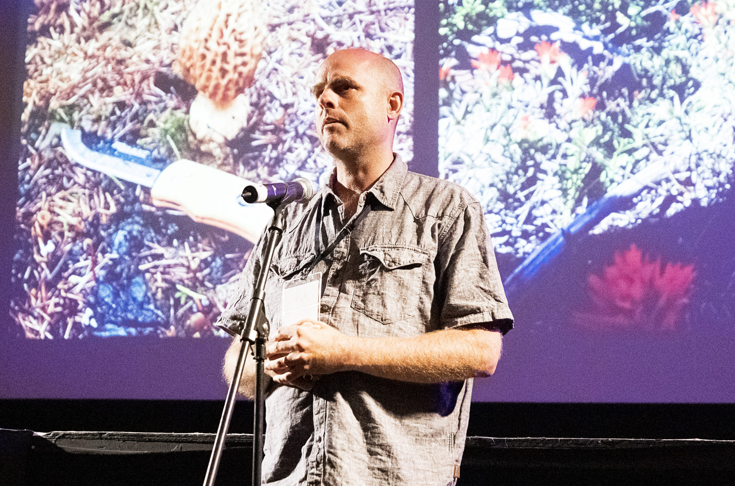 man with button down shirt and bald head standing in front a large movie screen talking in a microphone