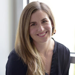 Molly Curley OBrien, with long light brown hair in a black top, smiles while sitting by a window, perhaps reviewing a rebuild permit.