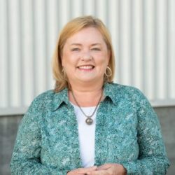 Pamela Van Halsema, a smiling woman with blonde hair wearing a green patterned shirt and necklace, stands confidently in front of a striped wall, ready to discuss the rebuild permit process.
