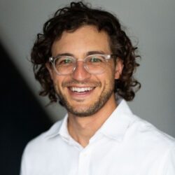 Smiling man with curly hair and glasses, wearing a white shirt, against a blurred background—Mikey Latner appears ready to discuss your rebuild permit needs.